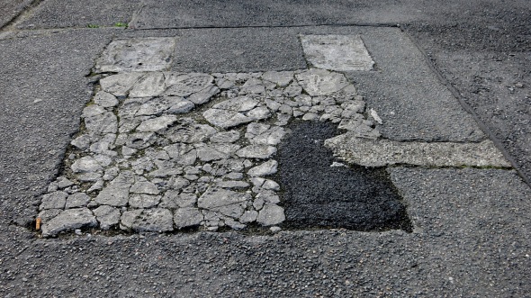 Kleines Stück eines alten asphaltierten Bahnsteigs, an einer Stelle mit Beton geflickt, der seinerseits an zwei Stellen wieder mit Asphalt geflickt ist.