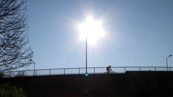 Blick gegen die Sonne auf eine Brücke; Brücke, Geländer und Fahrradfahrer als Silhouette vor dem blauen Himmel; am oberen Ende eines Laternenpfahls die Sonne als überbelichtetes sternförmiges Gebilde; am Fuß des Laternenpfahls ein leuchtend blauer Punkt - irgendein Artefakt oder eine Spiegelung im Objektiv