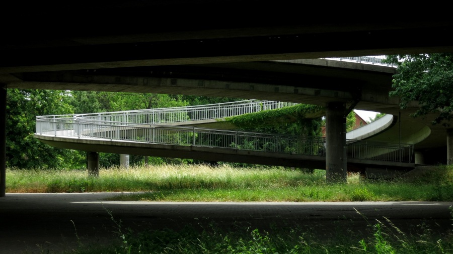 Kreisförmig gebauter Fußweg, der auf eine große Straßenbrücke führt, gesehen von der Seite; am oberen Bildrand die Unterseite der Brücke, am unteren Bildrand gepflasterte Oberfläche; der Fußweg eingerahmt von Gras und Bäumen