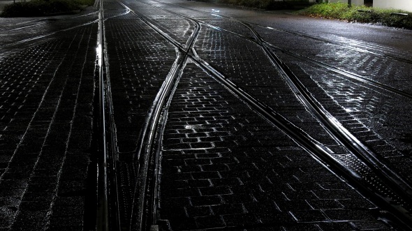 Schwarze, quadratische Pflastersteine, die Fugen mit Asphalt verklebt, und Straßenbahnschienen mit Weichen im Gegenlicht von Straßenlaternen.