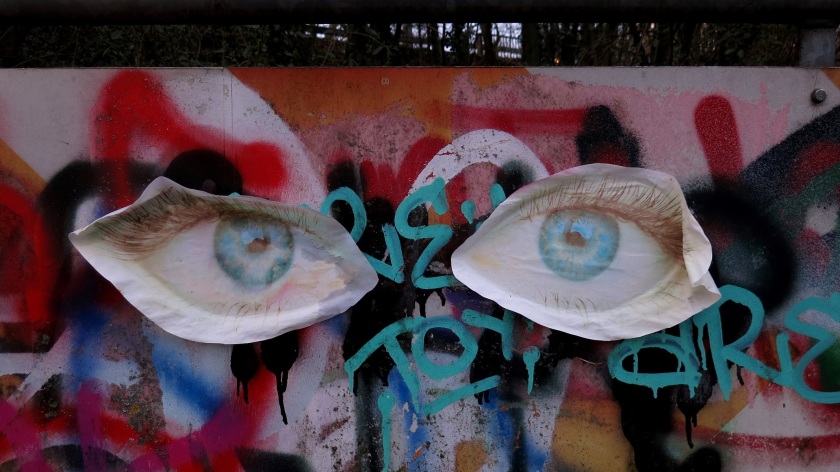 Mit Grafitti bedeckte, eigentlich rotweiße Warntafel am Rand eines Fuß- und Radwegs im Park; zwei etwa handgroße Augen sind auf Papier gedruckt, ausgeschnitten und aufgeklebt.
