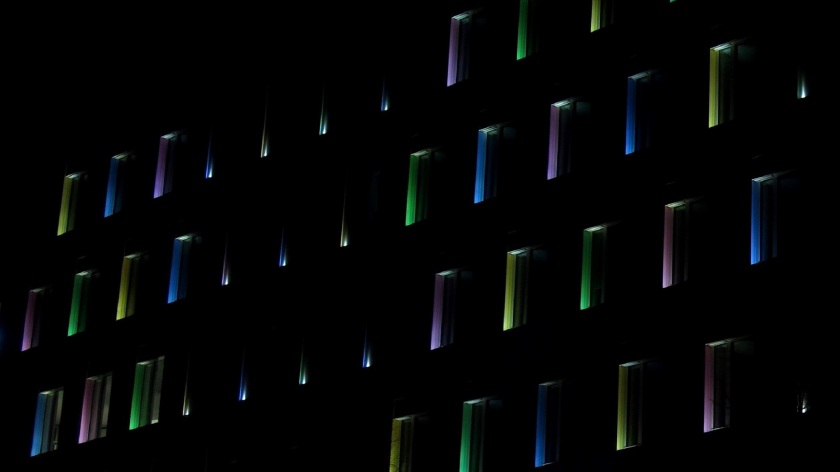 Bei Dunkelheit fotografierte Fassade eines Hauses mit gleichförmigen Fensterreihen, deren Rahmen von innen farbig beleuchtet sind, grün, gelb, blau, lila wechseln sich in loser Folge ab.