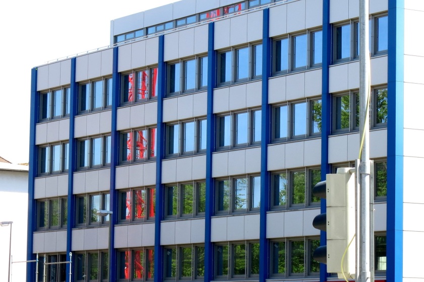 Fassade eines Geschäftsgebäudes mit blauen Strukturlementen und Fensterzeilen, in denen sich der blaue Himmel und ein roter Kran von der Baustelle nebenan spiegelt.