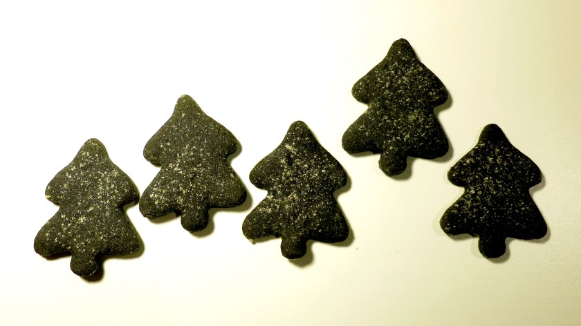 Fünf aus schwarzgefärbtem Teig hergestellte Kekse in Form von stilisierten Tannenbäumen, mit silbrigem Puder bestäubt, vor weißem Hintergrund