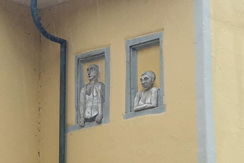Wand eines älteren Hauses mit zwei Blindfenstern auf halber Höhe; in den Fenstern sind ein Mann und eine Frau als Relief abgebildet.